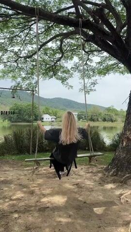 A woman with long, blonde hair is sitting on a rope swing under a large tree. The swing faces a calm lake reflecting the sky. Two white buildings are visible on the other side of the lake, backed by lush green hills. The ground under the tree is covered with dry leaves. The woman is wearing a dark shirt and black pants. She has her hands up, holding the ropes of the swing. The scene appears peaceful and serene.