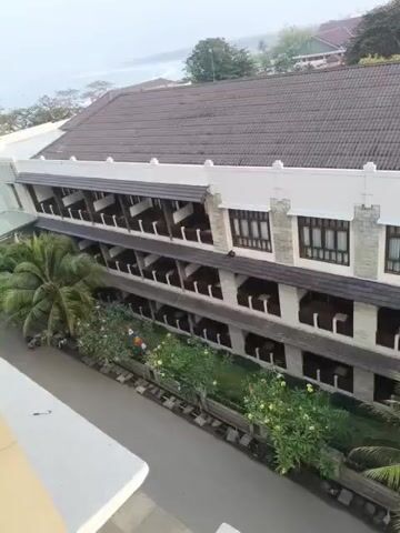 The video shows an aerial view of a hotel building. The building has multiple floors with balconies and windows. There are palm trees and other greenery surrounding the building. In the background, there is a body of water, likely the ocean, and other buildings. The sky is overcast. There are no people visible in the video.