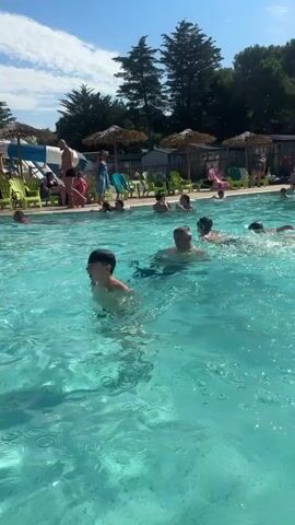 A group of people are swimming in a bright blue swimming pool on a sunny day. In the foreground, a man with a bald head and sunglasses is swimming towards the camera. To his left, a young boy with short dark hair is also swimming. Further back, other swimmers are visible, some wearing red flotation devices. The pool is surrounded by lounge chairs and umbrellas, and trees and mobile homes can be seen in the background. The water is clear and appears to be well-maintained. The overall scene suggests a recreational activity in a warm, outdoor setting.