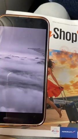 The video shows a close-up view of a smartphone displaying a photo of an airplane wing extending over a cloudy sky with mountains visible in the distance. The phone is placed on top of an open magazine. The magazine cover features a photo of a woman in an orange dress running with a suitcase in front of an airplane and airport terminal. The magazine has the word "Shop" in large letters and also displays logos for "Aeroflot" and "Aeromar". The lighting appears to be natural, and the overall scene suggests travel or a travel-related theme.