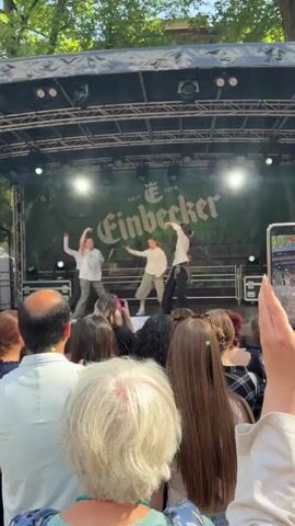 A group of four young people are performing a choreographed dance on an outdoor stage. The stage has a green banner behind them with the word "Einbecker" in large white lettering. The performers are dressed in casual, loose-fitting clothing. They are facing a crowd of people, some of whom are holding up phones to record the performance. The environment appears to be a park or outdoor festival, with trees visible above the stage. The lighting is bright, suggesting it is daytime. One person in the foreground is holding up a phone, capturing a video of the performance.