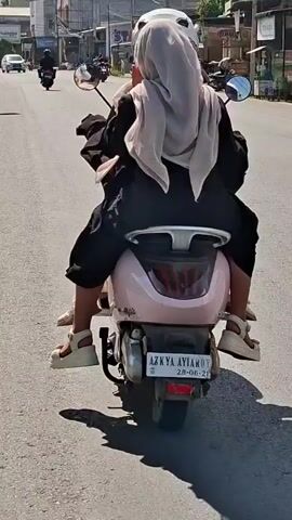 TWO PEOPLE, DRESSED IN CASUAL CLOTHING, ARE RIDING A LIGHT PINK MOTORCYCLE ON A PAVED ROAD. THE PERSON IN THE FRONT IS WEARING A WHITE HELMET AND DARK CLOTHING. THE PERSON IN THE BACK IS WEARING A LIGHT-COLORED HIJAB AND DARK CLOTHING, AND APPEARS TO BE WEARING WHITE SANDALS. THE MOTORCYCLE HAS A LICENSE PLATE THAT READS "AZKYA AYIAMON 28-06-21". THE BACKGROUND SHOWS A STREET WITH BUILDINGS AND OTHER VEHICLES, INCLUDING A WHITE CAR AND ANOTHER MOTORCYCLE IN THE DISTANCE. THE SUN IS SHINING, CASTING SHADOWS ON THE ROAD.