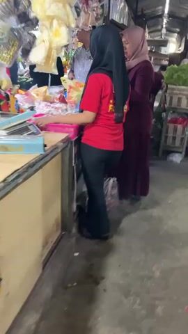 The video shows the interior of a market stall. Two women are visible. The woman in the foreground wears a red shirt and black pants, and her back is to the camera. The second woman, standing behind and to the right of the first, wears a maroon dress and a light pink headscarf. She is facing the first woman, and appears to be speaking to her. In front of the first woman is a stall counter with various items, including what appears to be food packaged in plastic. Behind the second woman, further market stalls are visible, stocked with produce like lettuce. The floor is concrete and appears worn.