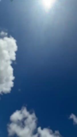 This is a vertical video of a bright blue sky with white clouds. The sun is visible in the upper right corner, casting a bright glare. The clouds are scattered throughout the sky, with a larger cloud formation on the left side and a smaller one near the bottom. The overall impression is of a clear and sunny day.