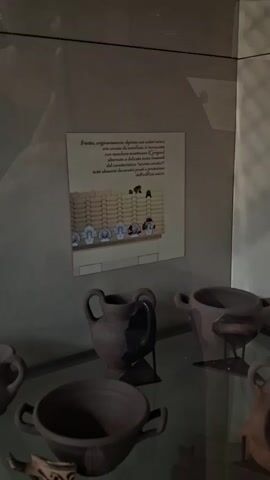 A digital photograph displays a museum exhibit featuring ancient pottery. In the foreground, several terracotta vessels with handles and wide openings are visible, arranged on a reflective surface, likely a glass shelf. The vessels are dark brown, some appearing intact and others with visible damage or missing pieces. Behind the pottery, on the wall, is a framed informational plaque. The plaque features an illustration of a tiled roof with decorative antefixes, which are ornamental architectural elements. These antefixes appear to be terracotta and depict faces, some stylized and some resembling masks. The illustration is accompanied by text in Italian, likely describing the architectural features or the origin of the artifacts. The lighting in the museum appears to be ambient, creating soft shadows and highlighting the objects. The overall impression is one of historical preservation and display.