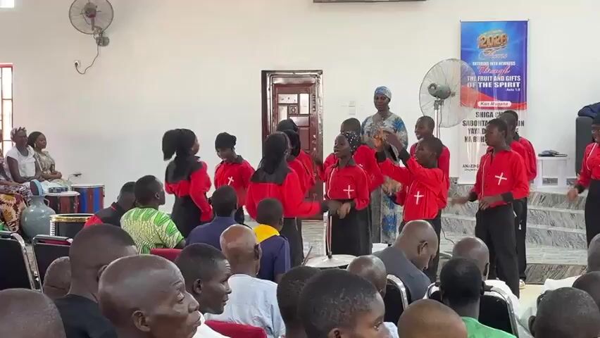 A group of people wearing red and black outfits are dancing and singing in front of an audience, as part of a performance. A banner behind them has the text "2023" and "THE FRUIT AND GIFTS OF THE SPIRIT". The audience is seated in front of the performers. The performance is taking place indoors in a well-lit room.