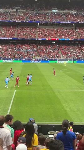 A soccer match is underway in a large stadium with many spectators. The field is green with white lines. Two teams are playing, one in red uniforms and the other in light blue uniforms. The players are running and kicking a soccer ball. A goalkeeper is in the net. The spectators are seated in the stands, wearing various colors. A large screen is visible above the field. The game appears to be in progress.