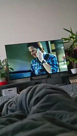 A television screen displays a scene from a show. On the left side of the screen, a man with long brown hair and a blue plaid shirt is on the phone. He is wearing a black watch on his left wrist. The background behind him is out of focus. The screen then transitions to a new scene. On the left, a woman with brown hair and a blue jacket is standing with her arms crossed. On the right, the man from the previous scene, now wearing a tan trench coat, is on the phone. Behind them is a sign that reads "Buster's Drive-In" and a field of green grass under a blue sky. The television is set on a black stand, and in front of it is a grey console with various objects, including a gray storage bin on the left and plants on the right. In the foreground, a person is covered by a gray blanket, partially obscuring the lower half of the television and the area below it.