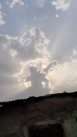 The video shows a cloudy sky with the sun peeking through a large cloud formation. The sunlight creates rays that shine down through the gaps in the clouds. At the bottom of the frame is the edge of a dark rooftop and the top of a building wall with some writing on it. The overall mood is somewhat dramatic with the contrast between the dark foreground and the bright sky.