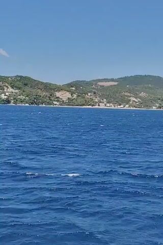 The video shows a wide shot of the blue ocean with small waves, indicating movement. In the background, a coastline with green hills and scattered buildings is visible under a clear blue sky. The camera appears to be moving, likely from a boat, as evidenced by the perspective of the water and the slightly blurry background. There are no people or specific objects clearly discernible in the frame.