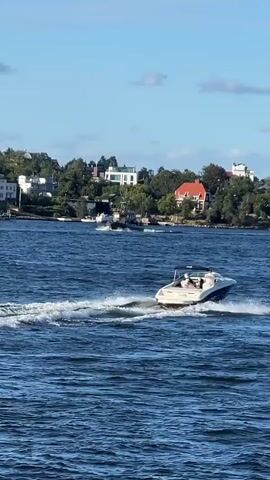 The video shows a bright, sunny day with a clear blue sky and some scattered white clouds over a body of water. The water is a deep blue and appears choppy with small waves. In the foreground, a white speedboat with a blue trim is moving at a moderate speed, leaving a white wake behind it. There are at least two people visible in the speedboat, one appears to be driving. In the background, across the water, there is a shoreline lined with lush green trees and several houses with red and white roofs. Further in the distance, another dark-colored boat is visible. The overall scene depicts a leisurely day on the water with boats navigating near a populated shoreline.