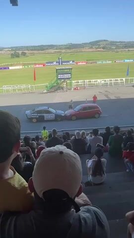The video shows a crowd of people sitting in bleacher seats, facing a racing track. Two cars, one black and one red, are positioned nose-to-nose on the track, with a person in a yellow vest standing between them. The black car appears to have some text or logos on its side. In the background, a green structure with stairs and a platform is visible, along with a grassy field and hills. The sky is clear and blue, indicating it is daytime. Some people in the crowd are holding up phones, presumably to record the event. The scene suggests an event involving a car demonstration or competition.