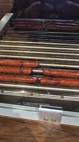 The video shows a close-up of a hot dog roller grill. There are two levels of rollers. The lower level has six hot dogs cooking on it, while the upper level has several more, which appear slightly darker in color. The rollers are slowly turning, rotating the hot dogs for even cooking. A person's reflection can be seen in the glass behind the grill. The grill is stainless steel with a wooden surface visible at the base. The brand name "HENDI" is visible on the front of the grill. The lighting appears to be indoor lighting.
