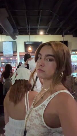 The video shows a young woman, likely in her 20s, standing in what seems to be a crowded bar or nightclub. She has shoulder-length brown hair and is wearing a white lace top and gold jewelry. Several people are around her, including a person with a white hoodie and a bandana, and another woman in a white top and red shorts. The bar area is visible in the background, with people standing near the counter, and TV screens on the wall. The lighting is somewhat dim with decorative light bulbs hanging from the ceiling. The woman in the foreground looks at the camera, smiles slightly, and then makes a "rock on" hand gesture. The overall environment appears lively and social.