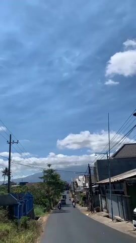 The video shows a slightly uphill, paved road in a village or small town. The sky is blue with scattered white clouds. On the left side of the road, there is a blue fence, greenery, and trees. On the right side, there are houses with tiled roofs and several utility poles with tangled wires. Several motorcycles are visible on the road, some with riders, and a few cars further up the road. In the distance, a mountain is faintly visible. The overall impression is a bright, sunny day in a residential area.