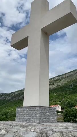 A large, monumental white concrete cross stands tall against a partly cloudy sky. The cross has a simple, blocky design, with a slightly textured surface. Its base is constructed from robust, rough-hewn grey stone blocks.

The image is captured from a low-angle perspective, looking up at the cross, which dominates the foreground. In the background, rugged green mountains and hills rise steeply, covered in scattered trees and shrubs, indicating a rocky landscape. Below the distant hills, a few small buildings with distinctive red-tiled roofs are visible, suggesting a small settlement or village.

The sky is a mix of bright blue and fluffy white clouds, suggesting daytime and fair weather. The ground in the immediate foreground appears to be paved with irregular grey stones. No people are visible in the scene. There is no clearly discernible text or inscription on the cross itself. The overall setting is natural and elevated, consistent with a landmark overlooking a landscape in Cijevna, Montenegro.