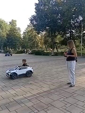 A young child sits in a white toy car. A woman stands to the right of the car, holding a remote control. The child looks to the left. The scene is outdoors on a paved area with trees and other people in the background. The toy car is positioned on the paved area. The woman is facing the toy car. The child's head is turned to the left. The toy car is white with black accents. The woman is wearing a dark top and light-colored pants. The child is wearing a dark top.