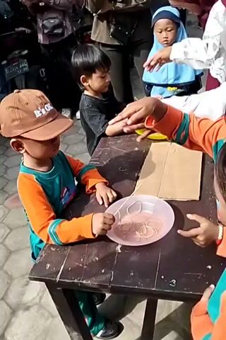 A group of young children are gathered around a wooden table outdoors, participating in what appears to be a game. One boy, wearing a brown cap and an orange and teal shirt, is intently focused on a pink plate filled with rubber bands, holding a stick. He is being instructed by an adult wearing a maroon hijab, who is pointing. Another boy in a black shirt sits at the table, also holding a stick. To the right, another child's hand reaches toward the table. In the background, more children and adults are visible. Some children are wearing blue hijabs. The ground is paved with hexagonal tiles. The overall setting seems to be an outdoor event or festival.