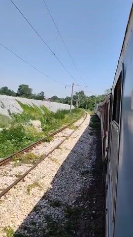 The video shows a view from a moving train. The train cars are visible on the right side of the frame, with their windows and a portion of their exterior. The train tracks run to the left, curving away into the distance. The landscape alongside the tracks consists of gravel, green vegetation, and a large pile of white debris or material, possibly from a construction site or quarry. Overhead, power lines and poles support the electrical supply for the train. The sky is clear and blue, indicating a sunny day. The camera perspective suggests the video was filmed from a window or the side of one of the train cars.