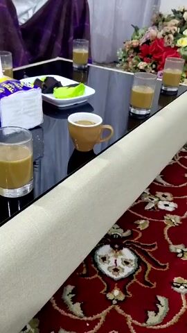The video presents a close-up view of a low black table covered with several glasses and a small brown cup, each containing a tan liquid. A small white dish with black and green sliced sweets is also on the table, alongside a pack of yellow and purple napkins. Part of a person in a white shirt and patterned tie is visible in the background, with their hands occasionally entering the frame. A cream-colored sofa or couch runs along the lower right edge of the frame. Below the sofa is a red rug with a floral pattern. Behind the table are floral arrangements and a person is partially obscured behind a purple draped cloth. The surface of the black table is reflective.