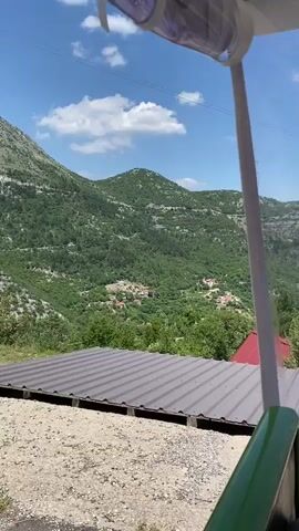 A video shows a scenic view of a mountainous area with lush green trees and a clear blue sky with some clouds. In the foreground, there are several small wooden cabins with red roofs. The cabins appear to be part of a resort or a small village nestled in the valley. The camera is positioned behind some sort of structure, possibly a canopy or a shelter, which partially obstructs the view. The video appears to be taken from a moving vehicle or a stationary vantage point overlooking the landscape.