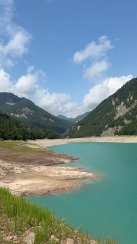 The video shows a turquoise lake surrounded by green mountains under a blue sky with scattered clouds. The water level appears to be low, exposing a sandy shoreline. In the foreground, there are rocks and green grass. The camera pans slowly across the landscape, offering a panoramic view of the serene natural environment. There are no people or animals visible in the video, and no discernible actions or interactions are taking place. The scene is peaceful and tranquil, highlighting the beauty of the mountainous terrain and the striking color of the lake.