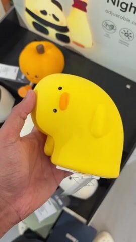 The media file is a short video clip featuring a close-up view of a person's hand holding a yellow, duck-shaped night light. The duck has small black eyes and an orange beak, giving it a cute and cartoonish appearance. The night light is soft to the touch, likely made of silicone, and its rounded form suggests it's designed to be child-friendly.

In the background, other items are visible, including a small orange pumpkin-shaped object and what appears to be packaging for other night lights, some with panda and lightbulb designs. A partially visible sign in the background contains the word "Night" and mentions "Adjustable".

The lighting suggests it's likely daytime, possibly indoors in a retail setting like a store or market, given the display of merchandise. The overall mood is cheerful and playful, conveyed by the cute design of the duck night light. No specific weather or time of day can be definitively determined, but the indoor setting is implied. The person's hand is the only human element visible.
