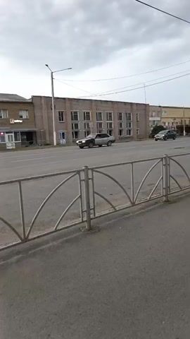 The video pans from left to right, showing a street scene with a cloudy sky. In the foreground, there is a sidewalk with a metal fence. Several cars are driving on the road. On the left side of the road, there is a building with multiple windows. Further down the road, there is another building with a lighter colored facade and windows. On the right side of the road, a group of people are walking on the sidewalk. There are trees and streetlights visible. The overall scene appears to be a moderately busy street in a town or city.
