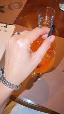 A close-up shot shows a hand holding a black straw in a glass of orange liquid with ice. The hand wears a silver bracelet and a ring. The background is blurred, with hints of a brown table and other glasses. The lighting is dim and warm.