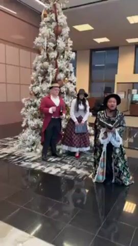 Four individuals in 19th-century attire are singing carols in front of a tall, decorated Christmas tree inside a building with a dark tiled floor.  The man on the left wears a red coat and holds what appears to be a tuning fork. The woman in the center wears a plaid skirt, a white vest, and a black hat. The man behind her is also in a top hat. The woman in the foreground is reading from a songbook and is dressed in a green and black floral gown with a white neck ruffle. Other people pass by.
