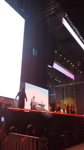 The video captures a large outdoor event at night, likely a public celebration. The primary focus is a massive LED screen displaying the text "FIESTAS PATRIAS en Maipú" in bold white letters, set against a background of red and blue, adorned with stylized stars and snowflakes. "Fiestas Patrias" translates to "National Holidays," and "Maipú" indicates the location of the event, which is in Chile.

Below the main text on the screen, an image of a person is visible, possibly an event host, performer, or public figure. In the background, a brightly lit stage can be seen with several individuals, appearing to be performing or presenting. The stage itself also features a circular screen with red and blue elements. The dark surroundings indicate nighttime, with stage lights illuminating the performers and the screen. The video is shot from an elevated perspective, looking down onto the event, and background audio includes music and crowd sounds, suggesting a lively atmosphere.