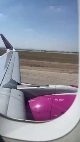This video captures a view from an airplane window during takeoff. The left side of the frame shows the wing of the aircraft, painted white with a purple stripe and branding. The engine, a prominent magenta color, is visible beneath the wing, with "NO STEP" clearly marked on its casing. The plane is on a runway, with the ground receding in the background. The airport tarmac and surrounding grassy areas are visible, along with distant buildings and a control tower. The sky is a clear blue with scattered clouds. The video captures the moment the plane lifts off the ground, as evidenced by the increasing distance between the aircraft and the runway.