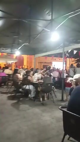 The video shows an outdoor gathering at night. There are several tables set up with people seated at them, appearing to be eating or socializing. A canopy covers the seating area, and string lights are visible. In the background, there is a building with illuminated windows and a stage area where performers or speakers might be. Pink and white pennant flags are strung overhead, suggesting a festive occasion. People of various ages are present, dressed in casual attire. The ground is paved with what appears to be cobblestones or rough stone. The overall atmosphere suggests a public event or outdoor restaurant setting.