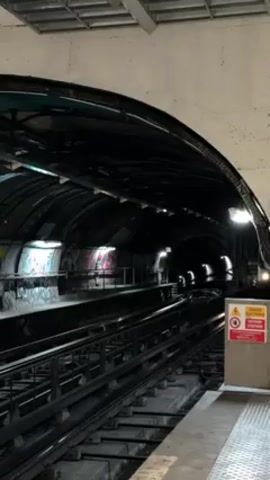 A subway train pulls into a station. The train is white with a black stripe and the acronym "RTM" written on the front. The station is underground with graffiti on the walls. There is a sign in front of the station warning of electric danger. The train is moving quickly as it pulls into the station. No people are visible.