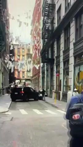 The video shows a street scene, likely in New York City. A black SUV is parked on the left side of the street. Overhead, white clothing hangs on lines strung between buildings, creating a visual spectacle. On the right side of the street, pedestrians are seen walking along the sidewalk and crossing the street. A building on the right is identified as "Manhattan Mini Storage" with the address "78A". There's a red fire hydrant near the entrance to the storage facility. The overall atmosphere seems urban and bustling.