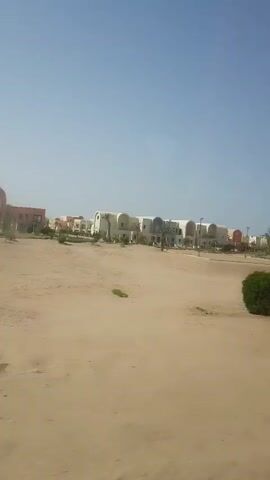 The video shows a view from a moving vehicle driving past a desert landscape with buildings in the background. The landscape is sandy with sparse vegetation consisting of shrubs and palm trees. The buildings in the background are mostly white with arched roofs and some are peach-colored. The sky is clear and blue. There are signs visible along the road. The vehicle appears to be moving at a moderate speed.