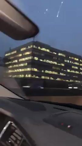 The video shows a view from inside a car driving on a road at dusk or early evening. Several large buildings with illuminated windows are visible in the background, along with trees and other vehicles on the road. The sky is overcast. The car appears to be moving in light traffic.