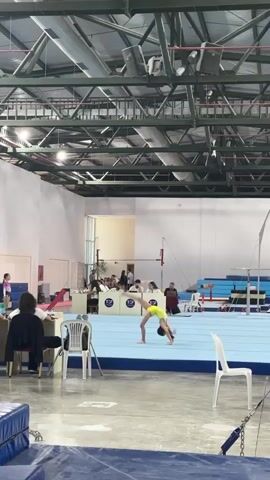 This media file captures a gymnastics competition taking place indoors. The main subject is a young female gymnast in a yellow leotard performing a routine on a blue mat. In the background, various gymnastics equipment is visible, including uneven bars and stacked mats. There are also spectators and judges seated at tables, suggesting an official event.

The setting appears to be a large gymnasium with high ceilings, exposed metal beams, and industrial lighting. White walls and a grey floor further contribute to the functional aesthetic of the space. A white chair is prominently placed in the foreground, and another is near the gymnast.

The gymnast is in the midst of a dynamic movement, possibly a leap or a split, demonstrating skill and agility. Her pose suggests focus and athletic prowess. The overall atmosphere seems to be one of focused competition.

There is visible text on the judges' tables, appearing as logos with the letters "T C" and possibly other symbols. The time of day is not explicitly indicated, but the indoor lighting suggests it could be any time. No specific weather conditions are discernible from the interior setting. The location context provided is Sancaktepe, Turkey.