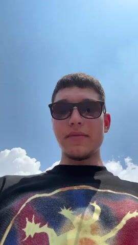 A young man wearing a black t-shirt with a graphic on it and brown sunglasses, films himself in front of a building with a Louis Vuitton store. Another young man walks in front of him. They are in a town center with many other people walking, and a few people riding bikes. The buildings are painted in various shades of yellow, peach, and orange. The sky is blue with white clouds.