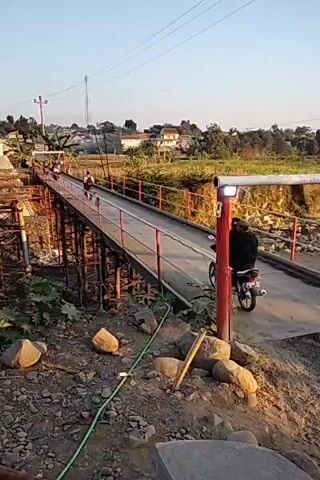 The video shows a narrow footbridge over a dry riverbed. A motorcycle with a rider wearing a dark jacket and pants is crossing the bridge. There are other people on the bridge in the distance. The bridge is supported by metal scaffolding. The surrounding area is rocky and overgrown with vegetation. In the background, there are houses and trees. The sky is clear and the lighting suggests it is late afternoon.