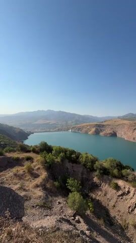 The video shows a panoramic view of a large, turquoise reservoir surrounded by arid, mountainous terrain. The sky is clear and blue, indicating a sunny day. In the foreground, there is a dry, rocky slope with sparse vegetation, including some green shrubs and trees. The reservoir dominates the middle ground, with its expansive water surface reflecting the clear sky. On the far side of the reservoir, a dam is visible, and beyond that, a range of hazy mountains stretches across the horizon. There are no people or visible man-made objects in the immediate foreground. The perspective suggests the video was taken from a high vantage point overlooking the landscape. The overall impression is one of vast, natural beauty and a sense of remoteness.