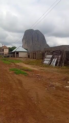 The video shows a dirt road in front of a large rock formation. There are several buildings and structures visible, including a small house with a metal roof, a partially constructed building with cinder blocks, and some stacked metal frames and panels. A few people can be seen near the house. The sky is overcast with clouds. A vehicle is parked on the left side of the road.