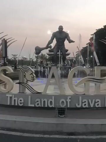 The video displays a large, stylized sign in the foreground that reads "SRAGEN The Land of Java." The sign features prominent, white, three-dimensional letters, with the letter "S" on the left incorporating a stylized depiction of a human profile with long, wavy hair. Behind the sign, a statue of a muscular, muscular male figure is visible, holding what appears to be a large club or hammer. The statue stands in front of a fountain with multiple water jets, some of which are illuminated with blue light. Several Indonesian flags, red and white, are planted on poles to the right of the statue. The background is a hazy sky with a soft, diffused light, suggesting either dawn or dusk. The overall setting appears to be an outdoor public space, possibly a roundabout or plaza, with some vegetation and indistinct structures in the distance. The video is a static shot, with no discernible movement or action beyond the water from the fountain.