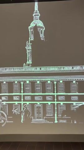 The video displays a projected animation of Independence Hall. The animation features a stylized, glowing green outline of the building, including its clock tower. In the foreground, two figures dressed in 18th-century attire are depicted. One figure, wearing a hat and coat, appears to be speaking to the other, who is holding a scroll or paper. A horse-drawn carriage is partially visible on the left side of the frame. The animation includes subtle movements, such as the hands on the clock tower ticking. The projection is cast onto a wall, with the bottom edge of the wall visible in the foreground. The overall impression is a historical reenactment or educational presentation.