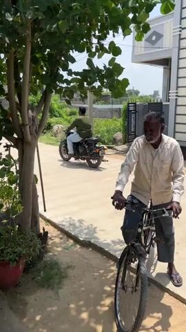 A man is shown from the waist down on the right side of the frame, sitting on a bicycle. He is wearing a light-colored shirt and blue jeans. The bicycle is black. In the background, a paved pathway extends into the distance. A tree with green leaves is on the left side of the frame, with a red potted plant beneath it. A motorcycle is parked in the background. The setting appears to be outdoors, with greenery and buildings visible in the distance.