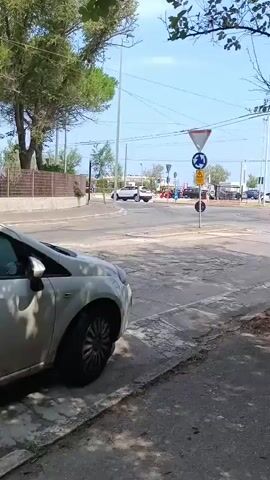 The video shows a street scene with a roundabout in the background. There are several cars visible, as well as traffic signs. A tree is in the foreground, partially obscuring the view. The sky is blue and it appears to be daytime. There is a white car parked on the left side of the frame.