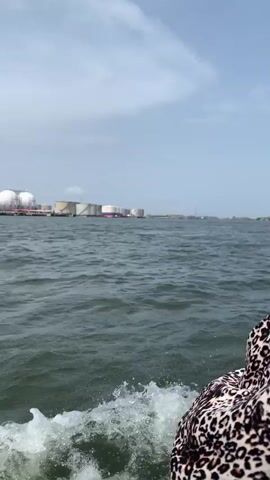 The video shows a body of water with gentle waves, likely an ocean or large bay. In the distance, a shoreline is visible with large industrial storage tanks. Some of the tanks are spherical and white, labeled "Rainoil." Other tanks are cylindrical. A small amount of what appears to be smoke or steam is rising from one of the tank areas. The camera is positioned on a boat moving through the water, with a small portion of a person's leopard print clothing visible in the lower right corner of the frame. The sky is overcast with a mix of white and gray clouds.