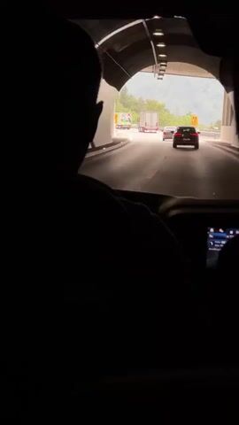 The video shows the interior of a vehicle, looking forward through the windshield and out of the back window. The perspective is from behind the driver, who is silhouetted and has their hands on the steering wheel. The vehicle is exiting a dark tunnel and entering a bright, outdoor environment. Outside the vehicle, there is a multi-lane highway with other vehicles, including cars and trucks. The highway is bordered by green hills and trees, and the sky appears overcast. The lighting transitions from dark to bright as the vehicle moves from the tunnel to the outside.