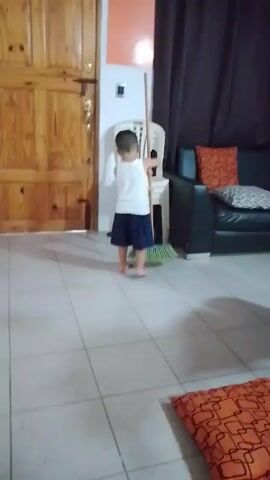 A young child is seen in a room. The child is wearing a white tank top and navy blue shorts. The child is near a broom that is leaning against a white plastic chair. A dark couch with pillows sits to the right. An orange pillow is in the bottom right corner of the screen. A wood paneled door is to the left. The child smiles and then walks out of the frame.