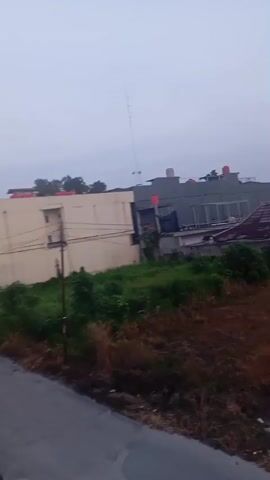 The video shows a blurry, overcast scene, possibly taken from a moving vehicle. The camera pans to the right, revealing several buildings, some with red roofs, partially obscured by trees and foliage. In the foreground, a paved road or path is visible. The sky is a uniform gray, suggesting a cloudy or misty day. The buildings appear to be residential or commercial, with some windows illuminated. There are no people visible in the footage.